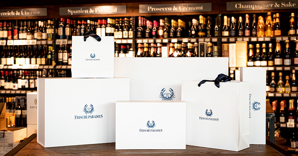 Verschiedene Frischeparadies Geschenkboxen in verschiedenen größen, sowie Geschenktüten. Im Hintergrund sind Weinregale zu sehen.